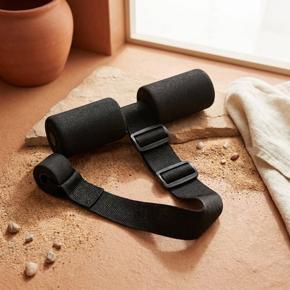 A pair of black foam roller massage bands with adjustable black plastic buckles, arranged on a textured terracotta surface with sand, a stone slab, and a beige cloth.